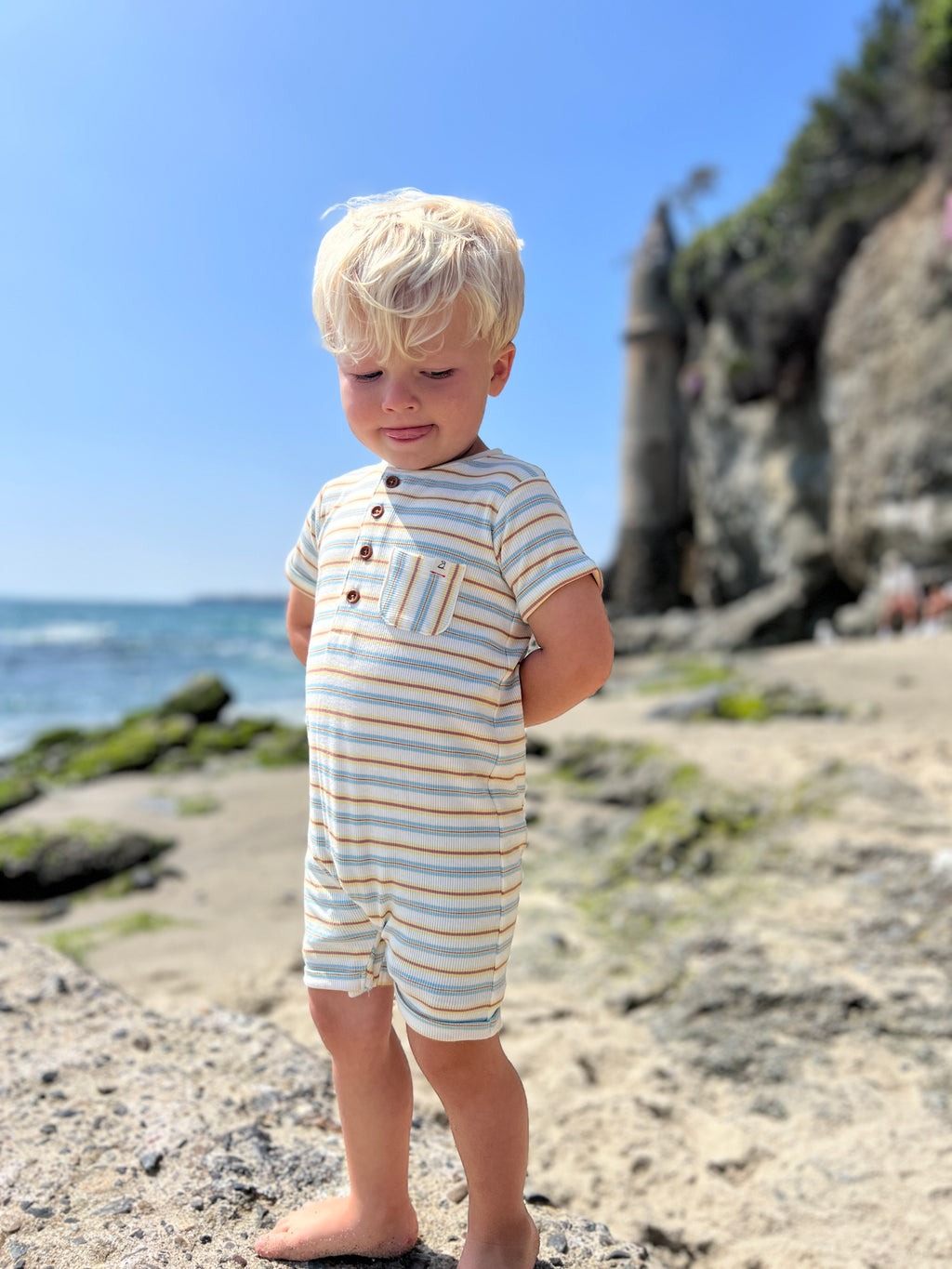 small blonde hair boy wearing our blue/lemon/white ribbed henley romper standing on rocks at the beach