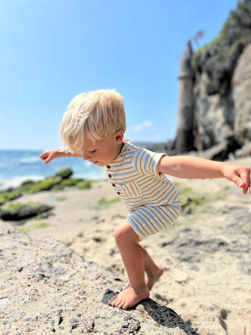 small blonde hair boy wearing our blue/lemon/white ribbed henley romper playing on rocks at the beach