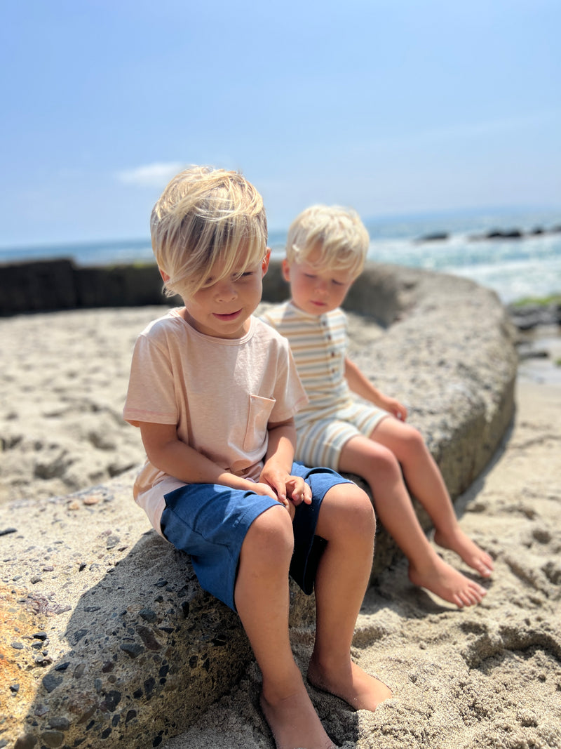 small blonde hair boy wearing our blue/lemon/white ribbed henley romper sitting on rocks at the beach with another blonde hair boy wearing our peach Josh tee and navy pique shorts.