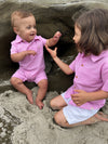 blonde hair blue eyed toddler wearing pink seersucker polo romper, with small boy with long brown hair wearing pink seersucker shirt and white crew shorts sitting on the sand at the beach in summer