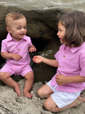 blonde hair blue eyed toddler wearing pink seersucker polo romper, with small boy with long brown hair wearing pink seersucker shirt and white crew shorts sitting on the sand at the beach in summer