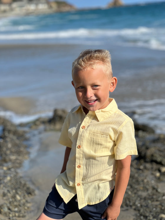 blonde hair boy wearing the lemon woven shirt and navy tiwill shorts by the sea at the beach.