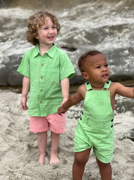 a boy with brown curly hair wearing our Lime Seersucker Woven Shirt and our pink pique shorts, holding hands with a small boy with. brown hair wearing our lime woven overalls., standing on the sand at the beach.