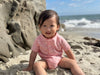 Child in a red striped jersey onesie sitting on a sandy beach with ocean waves in the background.