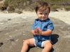 Child wearing Dark Chambray stripe woven onesie  sitting on a sandy beach.