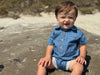 Child wearing Dark Chambray stripe woven onesie  sitting on a sandy beach.