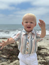 Child in a Chambray/Neon striped Woven Onesie and white pants  standing on a beach with ocean and sky in the background