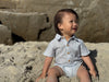 Child sitting on sand wearing a Chambray Textured woven onesie with large rocks in the background.