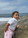 Child standing on a rock by the ocean in a white seersucker, woven onesie.