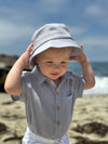 Child wearing a light grey bucket hat and a grey woven onesie on a beach with ocean and sky in the background