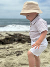 Child wearing a beige bucket hat and cream seersucker woven onesie. white shorts on a beach.