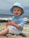 Child wearing a Pale blue bucket hat and chambray  stripe woven onesie, white shorts sitting on the sand.